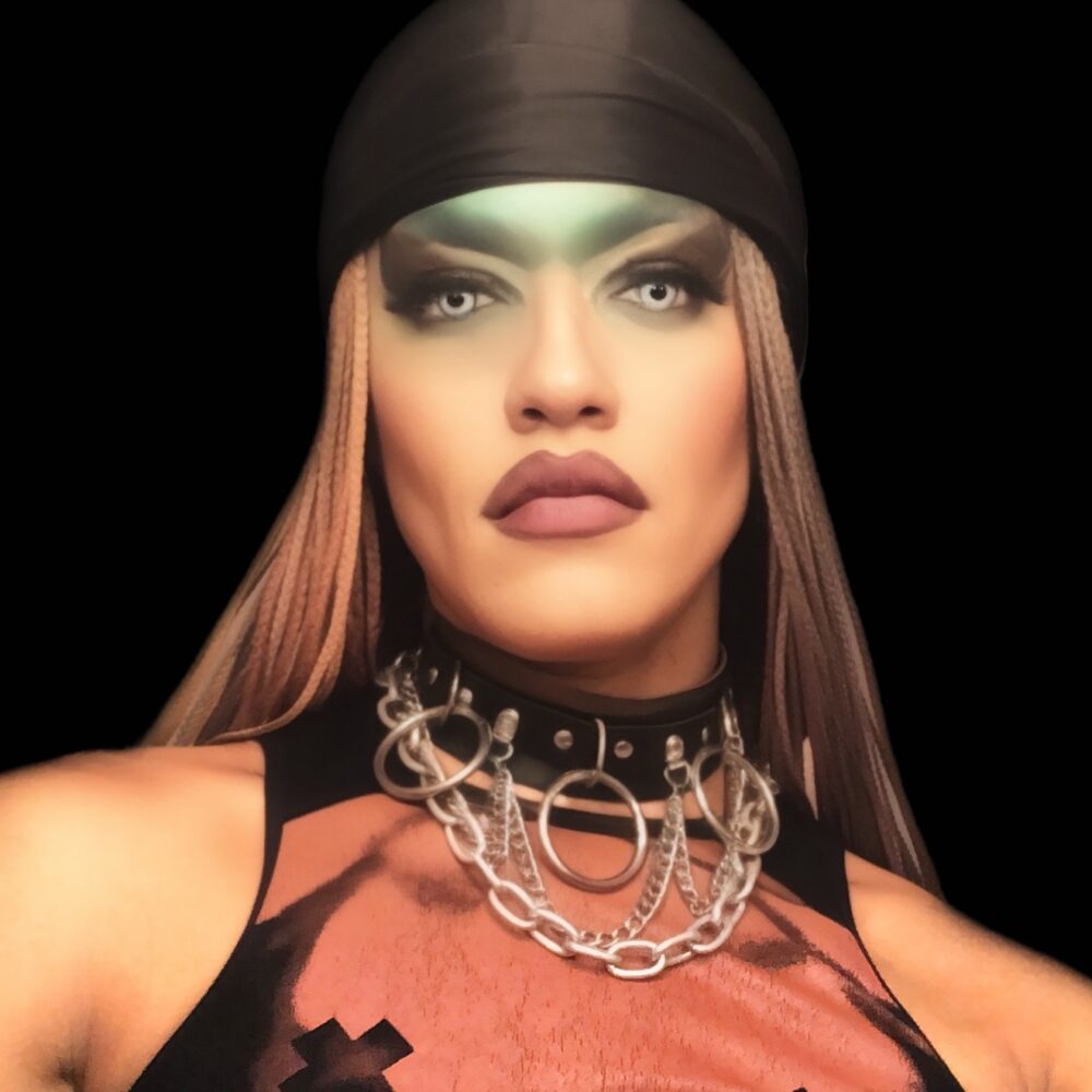 A photo of a person looking directly at the camera in front of a black background. They have long straight hair tucked behind their shoulders with a black bandana across their forehead. They are wearing striking green eye makeup and a black and orange vest top with a silver necklace.